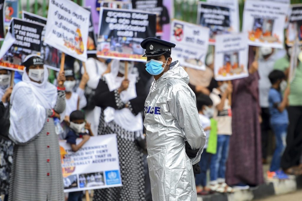 A Sri Lankan police officer stands guard as protesters hold placards during a demonstration against the government’s policy of forced cremations of Muslims who die of COVID-19 outside a cemetery in Colombo on Dec. 31, 2020. (Photo by Ishara S. Kodikara/AFP)